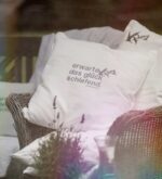 Weißes Kissen mit gesticktem Spruch auf Korbsessel im Outdoor-Bereich