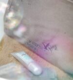 Makroaufnahme der Stickerei auf grauem Beautycase mit Pflegeprodukt im Vordergrund