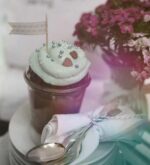 Kleiner Kuchen im Glas mit pastell-petrolfarbener Sahnehaube, Schokoherzen und Fähnchen auf Tellerstapel