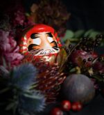 Roter Daruma zwischen dunklen Blumen, Beeren und Feigen; Symbol für Mut und Neustart, handbemalt, 9 cm