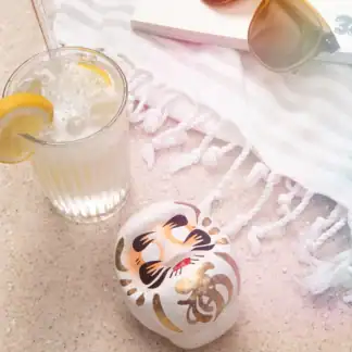 Weißer Daruma auf Sand neben sprudeligem Getränk, Sonnenbrille und Strandtuch